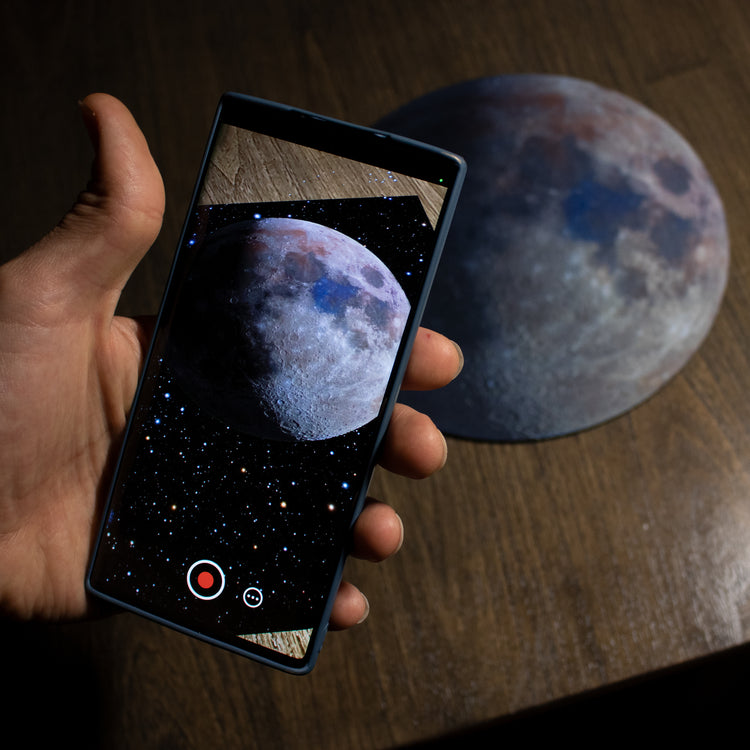Image has a round mousepad with moon art "Luna Bella" printed on it. Above the mousepad is a cellphone with camera pointed at mousepad demonstrating the Augmented Reality animation.